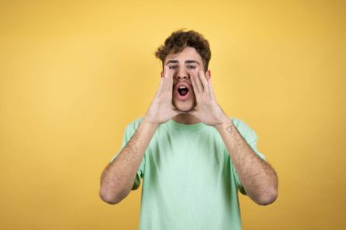 Sarı arka planda yeşil bir tişört giyen yakışıklı adam elleri ağızda bağırıyor ve bağırıyor.