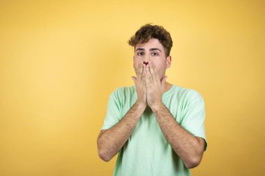 Sarı arka planda yeşil bir tişört giyen yakışıklı bir adam. Elleri ağzında ve şaşırmış.