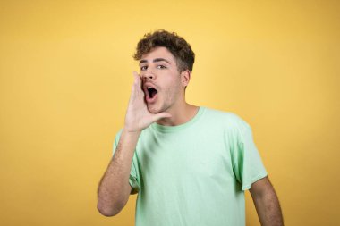 Sarı arka planda yeşil bir tişört giyen yakışıklı adam elleri ağızda bağırıyor ve bağırıyor.