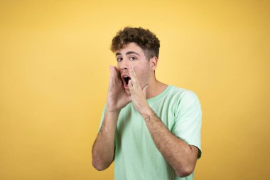 Sarı arka planda yeşil bir tişört giyen yakışıklı adam elleri ağızda bağırıyor ve bağırıyor.