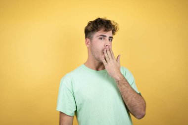 Sarı arka planda yeşil bir tişört giyen yakışıklı bir adam. Eli ağzının üstünde ve şaşırmış, yan bakıyor.