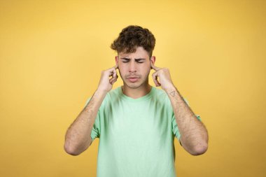 Sarı arka planda yeşil bir tişört giyen yakışıklı adam yüksek sesli müzik sesi için kulakları parmaklarıyla kapatıyor. Sağır kavramı.