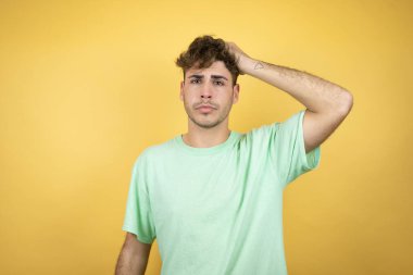 Sarı arka planda yeşil bir tişört giyen yakışıklı bir adam bir elini başının üstüne koyup sanki bir şey unutmuş gibi gülümsüyordu.