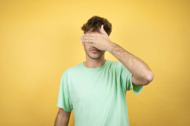 Sarı arka planda yeşil günlük tişört giyen yakışıklı bir adam. Ciddi ve elleriyle gözlerini kapatıyor.