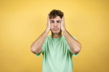 Sarı arka planda yeşil bir tişört giyen yakışıklı adam yorgun ve elleri başının üstündeyken sıkılmış görünüyor.