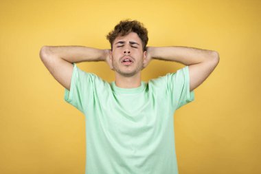 Sarı arka planda yeşil bir tişört giyen yakışıklı adam rahatlıyor ve esniyor, kolları ve elleri başının ve boynunun arkasında mutlu bir şekilde gülümsüyor.