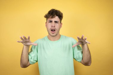 Sarı arka planda yeşil bir tişört giyen yakışıklı bir adam komik bir şekilde kedi gibi pençe hareketi yapıyor, agresif ve seksi bir ifade.