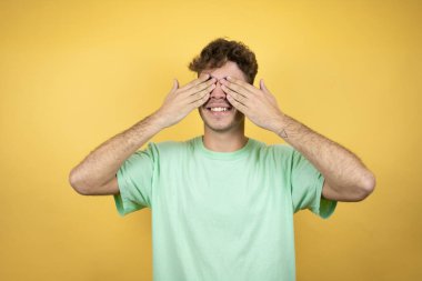 Sarı arka plan üzerine yeşil günlük tişört giyen yakışıklı bir adam. Gözleri güler yüzlü ve komik bir şekilde kapatıyor. Kör kavram.