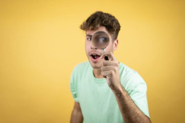 Sarı arka planda yeşil günlük tişört giyen yakışıklı bir adam büyüteçten bakarken şaşırdı.