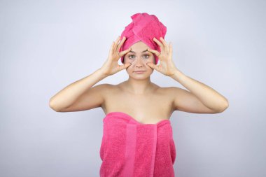Genç, güzel bir kadın duş havlusu takıyor. İzole edilmiş beyaz arka planda duruyor. Gözlerini parmaklarıyla açmaya çalışıyor.