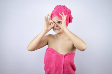 Genç, güzel bir kadın banyo üstüne duş havlusu giyiyor. İzole edilmiş beyaz arka planda dikiliyor. Gülümseyen yüzüyle, gözleriyle parmaklarının arasından bakıyor.