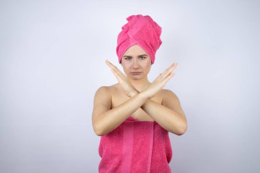Genç, güzel bir kadın banyo üstüne duş havlusu giyip izole edilmiş beyaz arka planda duruyor. Reddedilmiş yüz ifadesi kollarını kavuşturuyor. Negatif işaret veriyor.