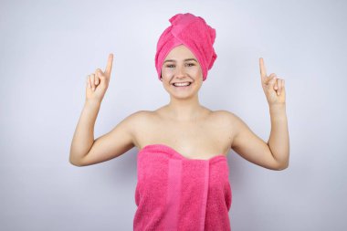 Genç, güzel bir kadın duş havlusu takıp izole edilmiş beyaz arka planda duruyor gülümsüyor, kameraya bakıyor ve parmaklarıyla işaret ediyor ve kollarını kaldırıyor.