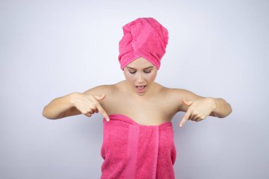 Genç, güzel bir kadın duş havlusu takıyor. İzole edilmiş beyaz arka planda duruyor. Aşağı bakıyor, parmaklarıyla işaret ediyor ve kollarını kaldırıyor.