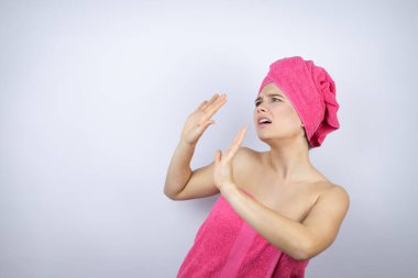 Genç, güzel bir kadın duş havlusu takıyor. İzole edilmiş beyaz arka planda duruyor. Kolları yukarıdan düşen bir şey gibi korkmuş.