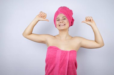 Genç, güzel bir kadın duş havlusu takıyor. İzole edilmiş beyaz arka planda duruyor. Yüzünde bir gülümsemeyle, gururlu ve mutlu bir şekilde kendini gösteriyor..