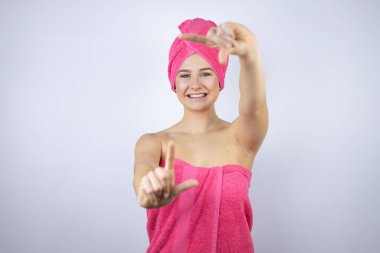 Genç, güzel bir kadın banyo üstüne duş havlusu giyiyor izole edilmiş beyaz arka planda duruyor gülümsüyor ve elleri ve parmakları mutlu yüzlü bir çerçeve yapıyor.
