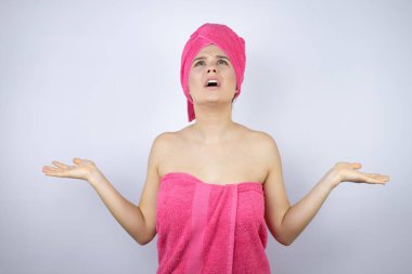 Banyodan sonra duş havlusu giyen genç ve güzel bir kadın izole edilmiş beyaz arka planda duruyor. Kolları ve elleri havada, kafası karışık bir yüz ifadesi var.