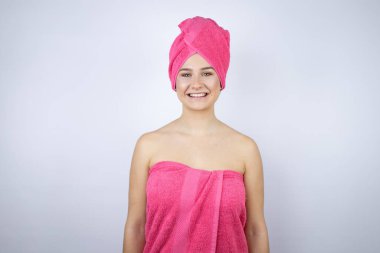 Genç, güzel bir kadın duş havlusu giyiyor. İzole edilmiş beyaz arka planda duruyor. Mutlu bir yüzü var.