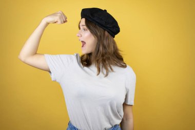 Fransız elbisesi giyen güzel beyaz kız izole edilmiş sarı arka planda bere takıyor ve gururla gülümseyen kol kaslarını gösteriyor.