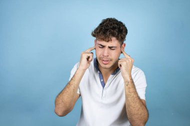 Mavi arka planda beyaz tişört giyen yakışıklı bir genç yüksek sesli müzik sesi için kulakları parmaklarıyla kapatıyor. Sağır kavramı.