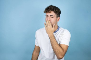 Mavi arka planda beyaz tişört giyen genç yakışıklı adam ağzıyla esnemekten sıkılmış. Huzursuz ve uykulu..
