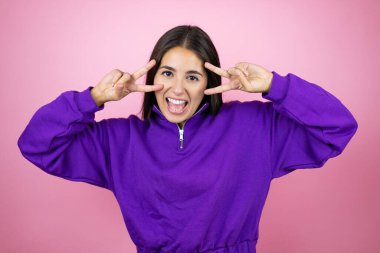 İnzivaya çekilmiş pembe arka planda eşofman üstü giyen genç ve güzel bir kadın barış sembolü yapıyor. Yüzünde parmaklar, gülümseyerek zaferi gösteriyor.