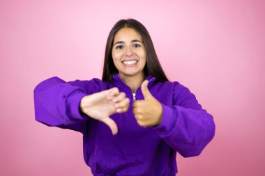 İzole edilmiş pembe arka plan üzerinde eşofman üstü giyen genç ve güzel bir kadın baş parmaklarını kaldırıyor, anlaşmazlık ve anlaşma ifadesi sergiliyor. Çılgın çatışma