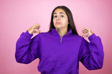 İzole edilmiş pembe arka plan üzerine kazak giyen genç ve güzel bir kadın tişörtünü başarılı bir ifadeyle tutuyor.