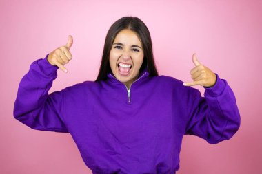 İzole edilmiş pembe arka planda kazak giyen genç ve güzel bir kadın çılgın bir ifadeyle el kaldırarak rock sembolü yapıyor.