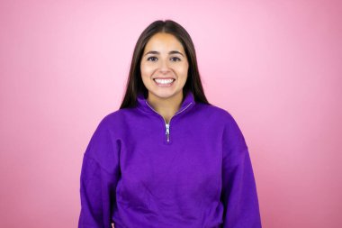 İnzivaya çekilmiş pembe arka planda eşofman üstü giyen genç ve güzel bir kadın. Mutlu bir yüzü olan ve kendine güvenen bir gülümsemeyle dişlerini gösteren.