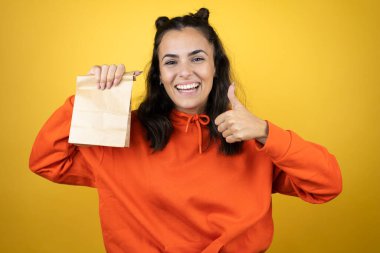 İzole edilmiş sarı arka plan üzerinde eşofman üstü giyen genç güzel bir kadın elinde bir kağıt torba başarı işareti, elinde olumlu bir jest, gülümseyen ve mutlu başparmaklar. Neşeli ifade ve kazanan jest.