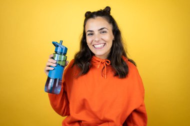 Tişört giyen genç bir kadın izole edilmiş sarı arka planda su şişesi tutuyor mutlu bir yüzü var ve gülümsüyor kendine güvenen bir gülümsemeyle dişlerini gösteriyor.