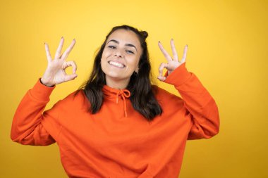 İzole edilmiş sarı arka plan üzerinde eşofman üstü giyen genç güzel bir kadın tamam işareti yapıyor parmaklarıyla ve gülümseyerek, mükemmel bir sembol.