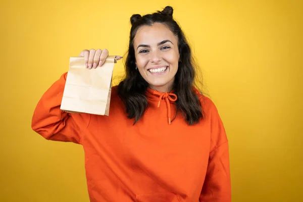 年轻美丽的女人穿着运动衫 披着孤立的黄色背景 微笑着 手里拿着纸袋 — 图库照片