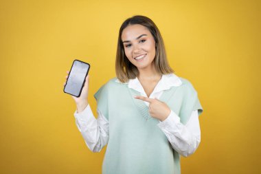 Sarı arka planda duran güzel genç bir kadın akıllı telefonu tutuyor ekranda gülümsüyor ve mutlu bir şekilde eli ve parmağı işaret ediyor.