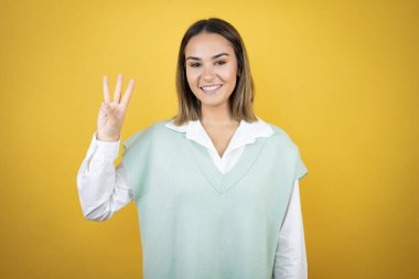 Sarı arka planda duran güzel genç bir kadın üç numarayı gösterip işaret ederken kendinden emin ve mutlu bir şekilde gülümsüyor.