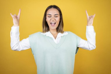 Sarı arka planda duran genç ve güzel bir kadın çılgın bir ifadeyle el kaldırarak rock sembolü yapıyor.