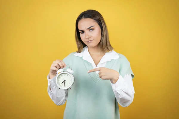 かなり若いです女性立ち上の黄色の背景深刻なホールディングとポインティングAクロック — ストック写真