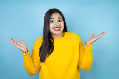 Sarı süveter giyen genç ve güzel bir kadın, izole edilmiş mavi arka plan bilgisiz ve elleri ve kolları havada, kafası karışmış bir ifade.