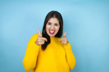 İzole edilmiş mavi arka planda sarı süveter giyen genç ve güzel bir kadın seni ve kamerayı işaret ediyor.