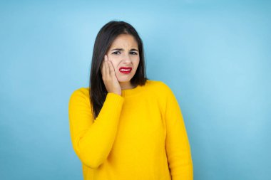 İzole edilmiş mavi arka planda sarı süveter giyen genç bir kadın diş ağrısı ya da diş ağrısı yüzünden ağız ve acı dolu bir ifadeyle dokunuyor.