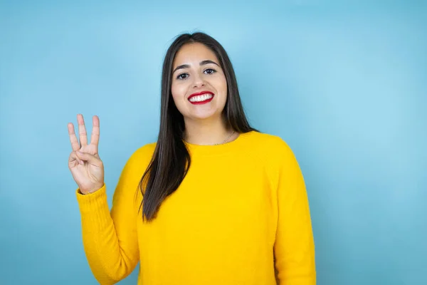 年轻漂亮的女人穿着黄色的毛衣 披着孤立的蓝色背景 带着自信和快乐的笑容 用手指着三号 — 图库照片