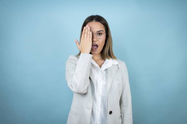 Genç iş kadını soyutlanmış mavi arka planda bir gözünü eliyle kapatıyor, yüzünde kendine güvenen bir gülümseme ve şaşırtıcı bir duygu var..