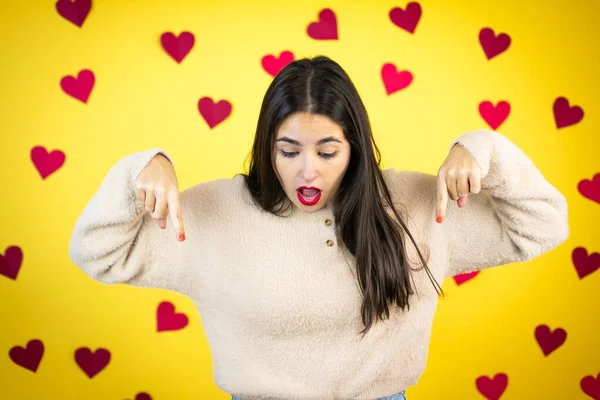 Young caucasian woman over yellow background with red hearts surprised, looking down and pointing down with fingers and raised arms