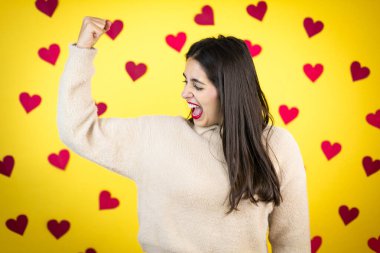 Sarı arka planda, kollarında gururla gülümseyen kasları olan beyaz bir kadın.