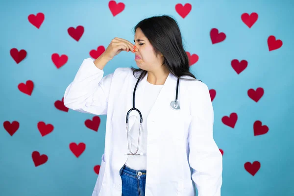 Mavi arka plan üzerinde steteskop ve tıbbi ceket giyen genç bir doktor. Kırmızı kalpler kötü ve iğrenç bir koku alıyor. Nefesini burnunda tutuyor.
