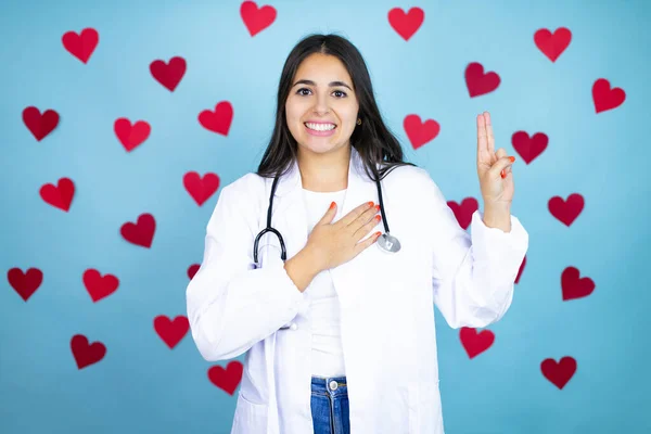 Young doctor woman wearing medical coat and stethoscope over blue background with red hearts smiling swearing with hand on chest and fingers up, making a loyalty promise oath