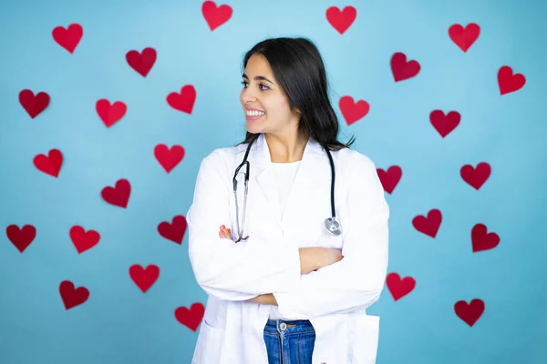 Young caucasian doctor woman wearing medical uniform and stethoscope over blue background with red hearts looking to side, relax profile pose with natural face and confident smile.