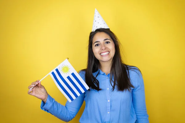 若いです白人女性の誕生日の帽子を身に着けています孤立した黄色の背景笑顔と保持ウルグアイの旗 — ストック写真